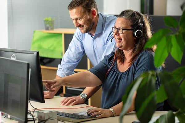Flytte telefonnummer til virksomhed To kolleger arbejder sammen ved en computer, hvor en medarbejder med headset betjener telefonisystemet til at flytte telefonummer til virksomhed